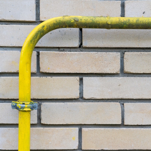 The yellow gas pipe is bent at right angles and is attached to a brick wall. The paint is covered with dirt in places. There are individual rustic bricks. Background. Texture.